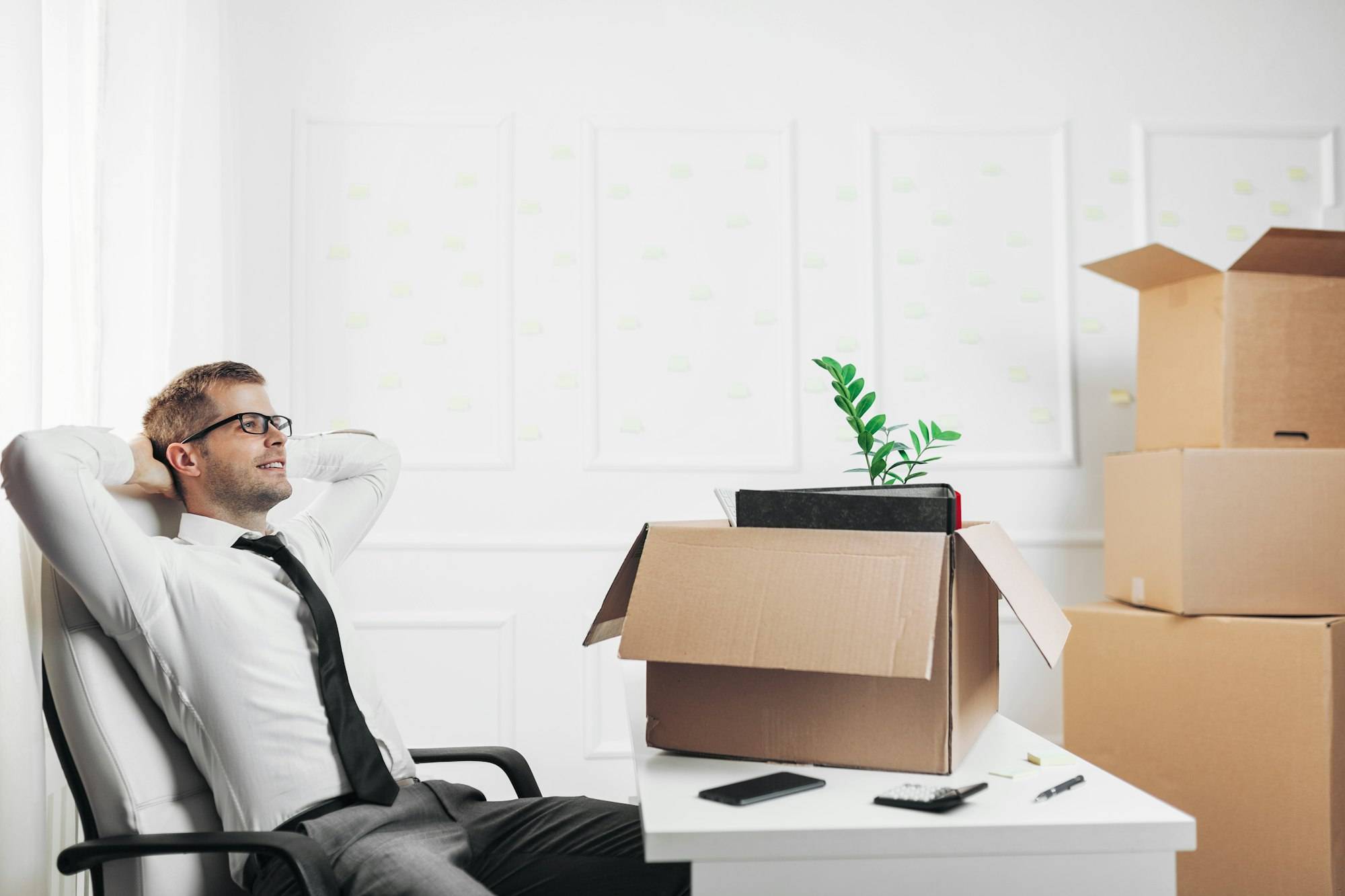 Homme d’affaires détendu dans un fauteuil, entouré de cartons et d’un vase avec une plante sur une table, ambiance déménagement.