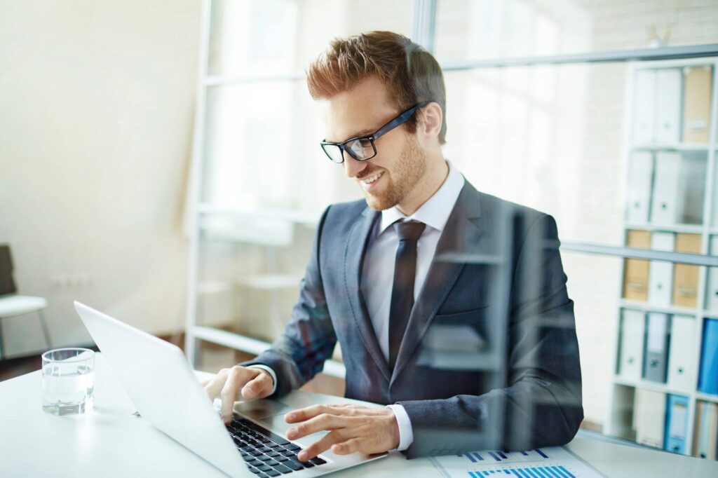 Homme d’affaires souriant et élégant en costume, travaillant sur un ordinateur portable dans un bureau lumineux.