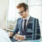 Homme d’affaires souriant et élégant en costume, travaillant sur un ordinateur portable dans un bureau lumineux.
