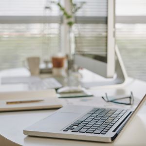 Bureau avec ordinateur portable ouvert sur un table, lunettes et documents, près d’une fenêtre lumineuse. Déménagement démarches à organiser.