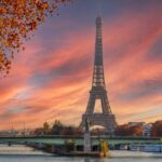 Vue de face de la tour Eiffel au coucher du soleil avec le fleuve et un pont au premier plan et des arbres aux couleurs