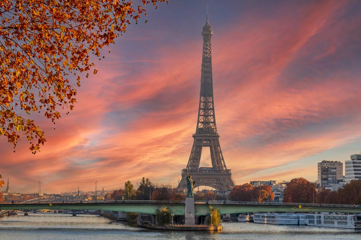 Vue de face de la tour Eiffel au coucher du soleil avec le fleuve et un pont au premier plan et des arbres aux couleurs